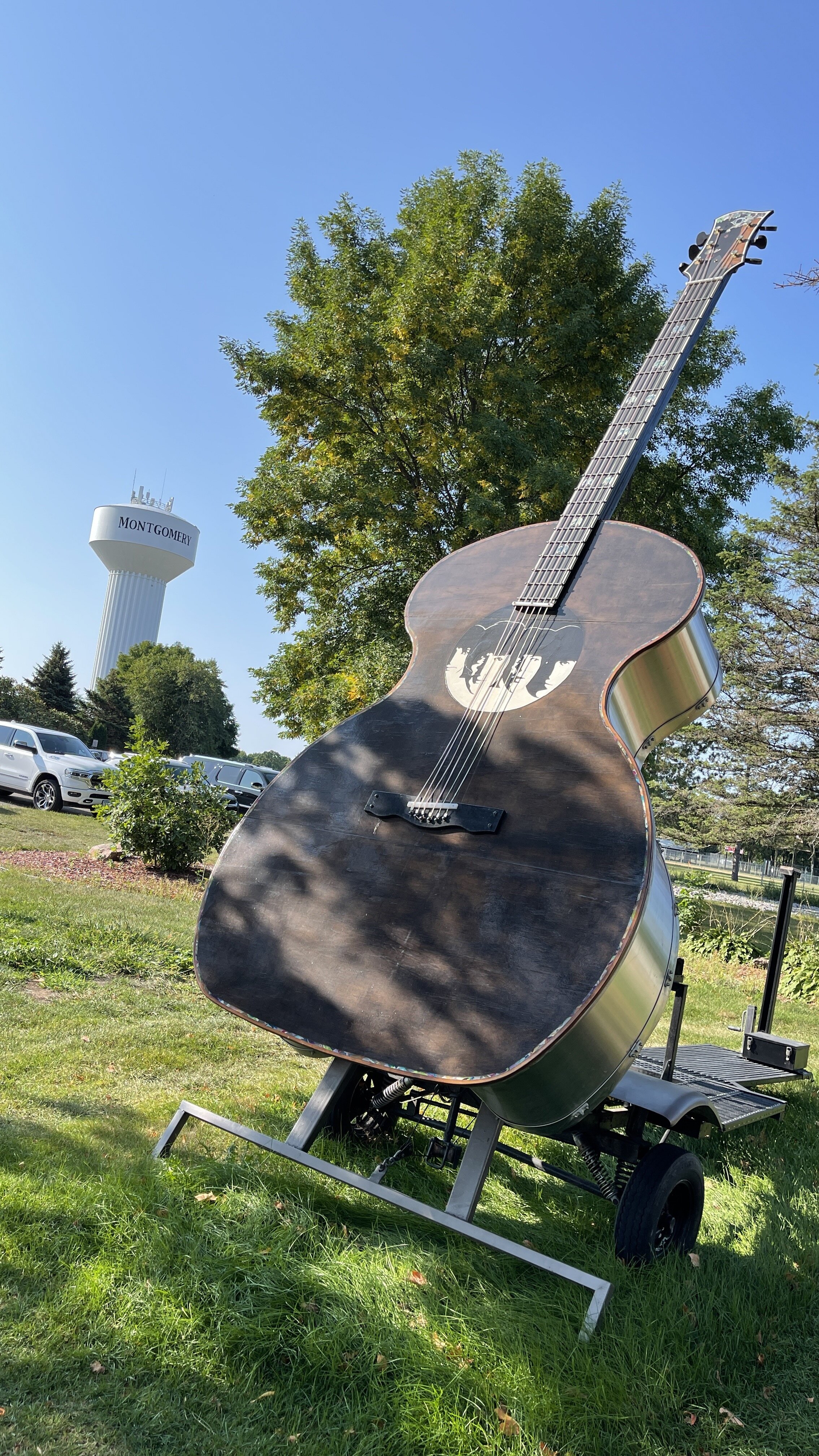 A giant acoustic guitar sculpture on a trailer on a lawn near parked cars, with the Montgomery water tower in the distance