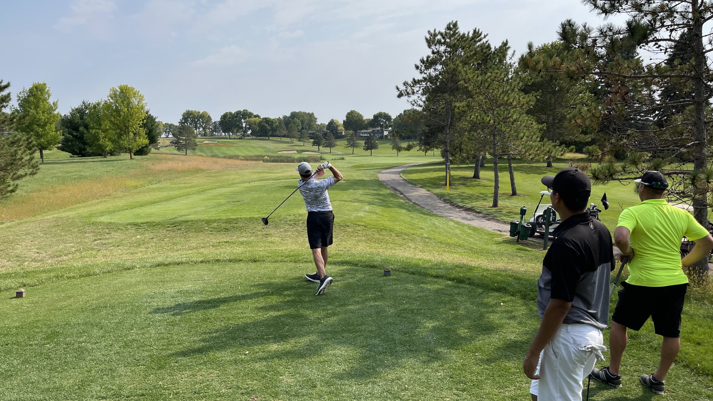 A golfer tees off as two playing partners wait beside the tee box, with golf carts parked along the path, at Montgomery National Golf Club