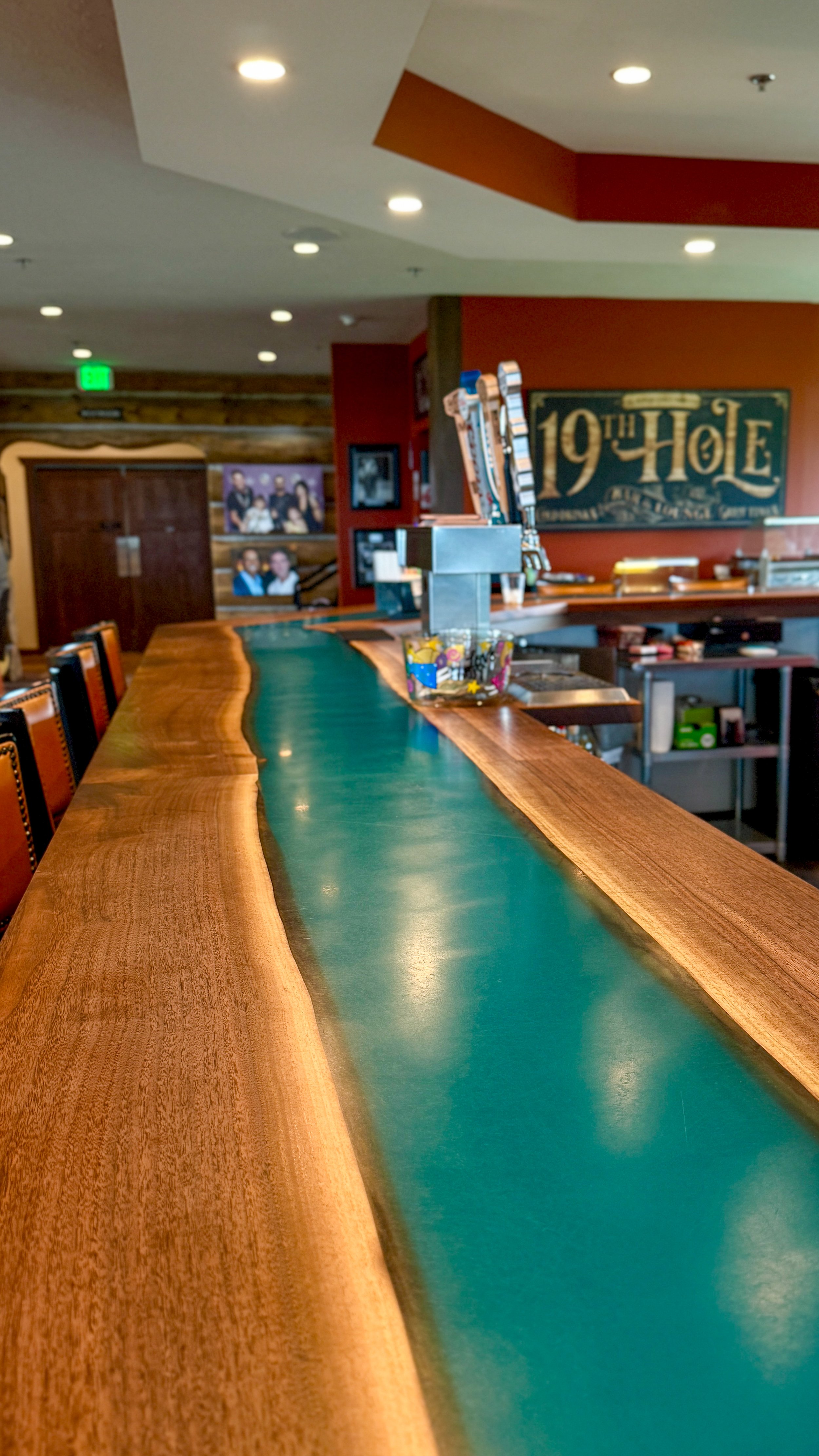 A long live-edge wood bar with a teal inlay, with a 19th Hole sign on the wall behind it, inside the Montgomery National clubhouse